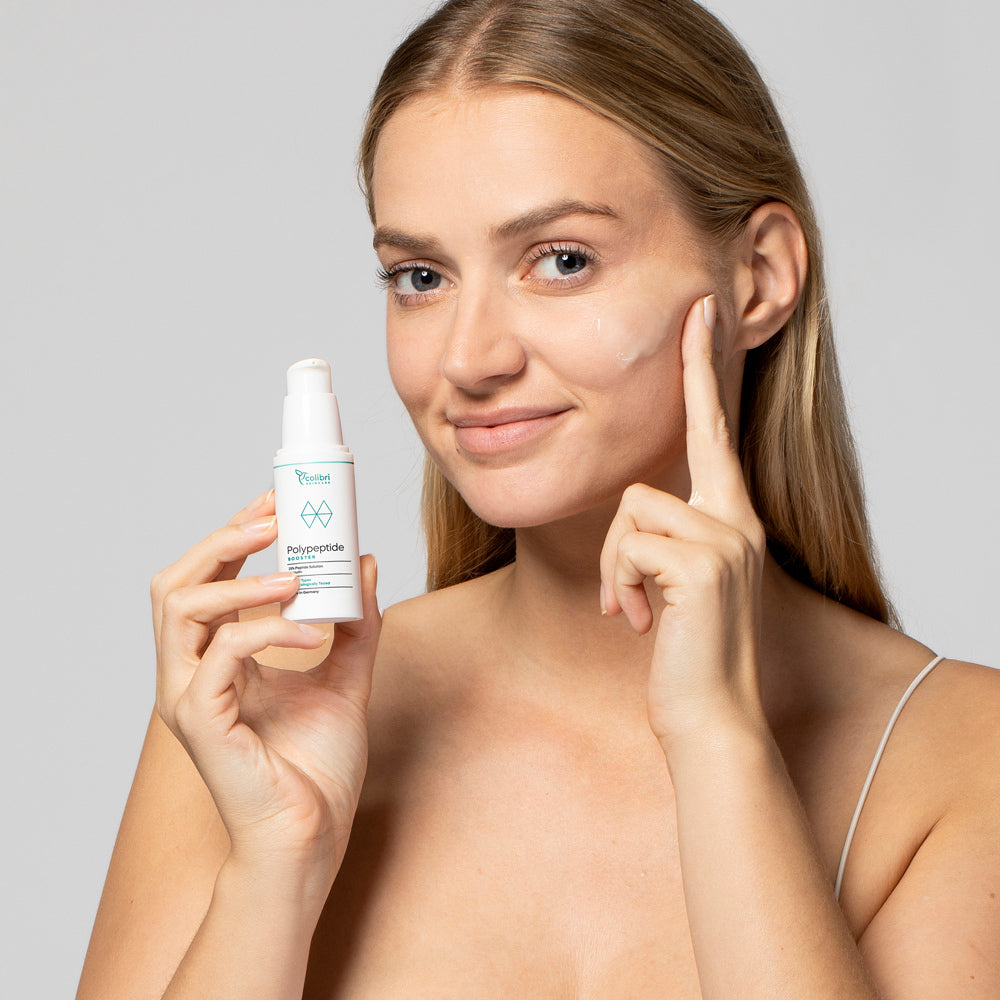 Eine Frau trägt Creme aus einem Spender auf ihre Wange auf und lächelt in die Kamera.