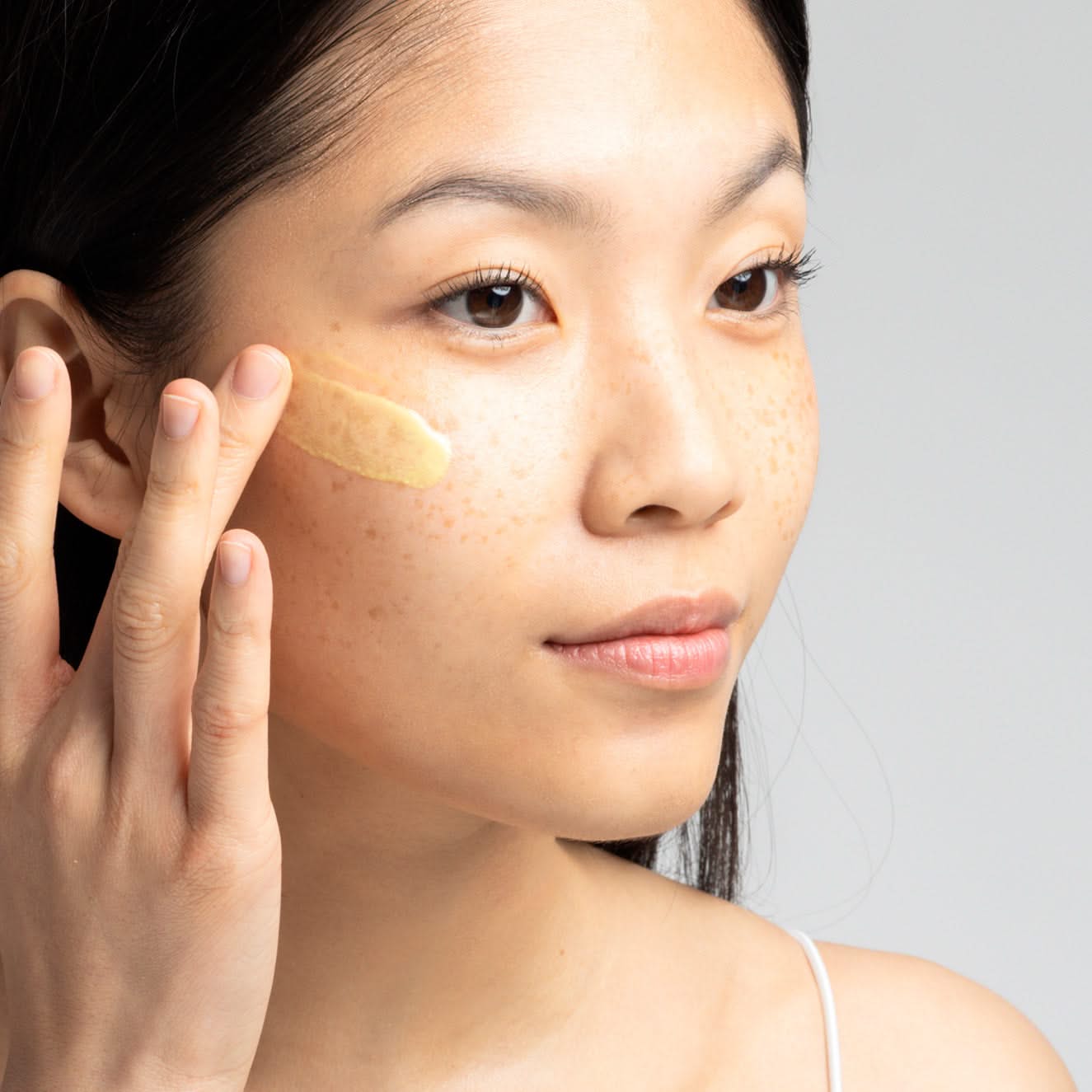 Frau trägt Hautpflegecreme auf die Wange auf, grauer Hintergrund.