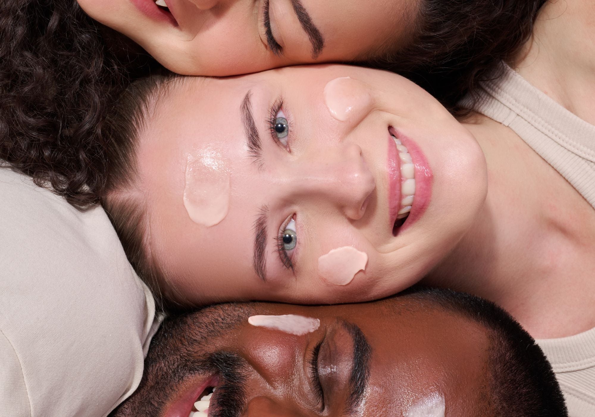 Drei lächelnde Personen, nebeneinander liegend, mit Gesichtsmasken auf den Wangen und Stirn.