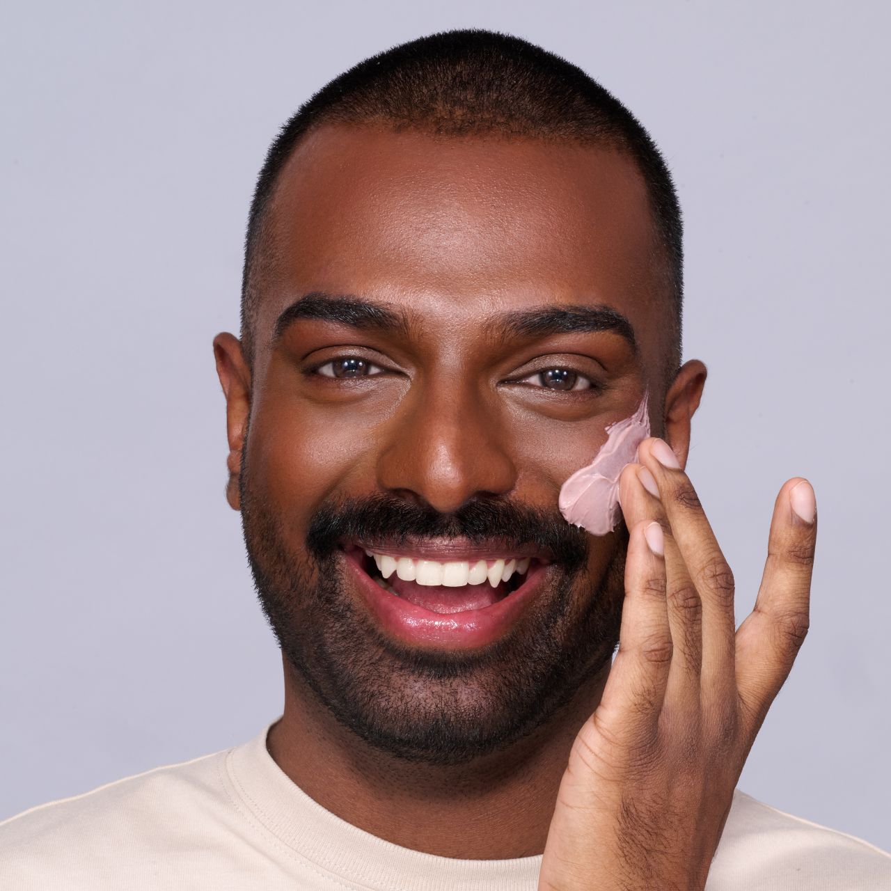 Ein lächelnder Mann trägt rosa Creme auf seine Wange auf und schaut direkt in die Kamera.