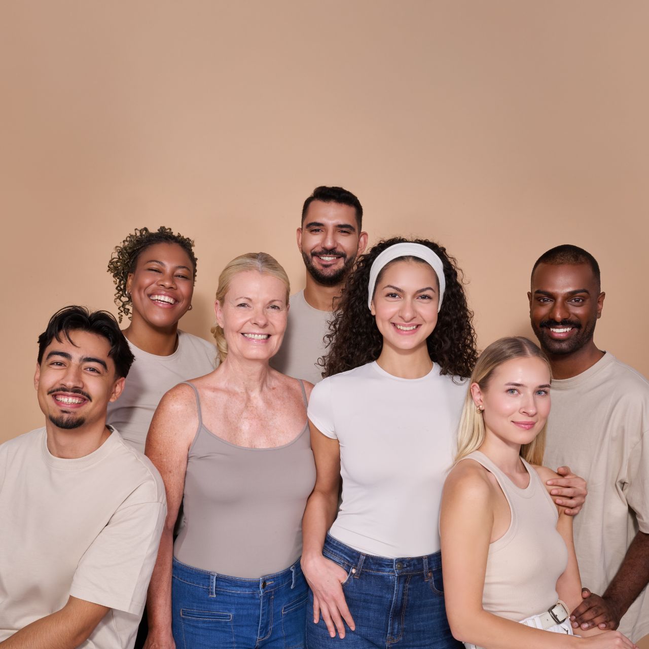 Sieben Personen in neutralen Tönen gekleidet lächeln gemeinsam vor einem beige Hintergrund in die Kamera.