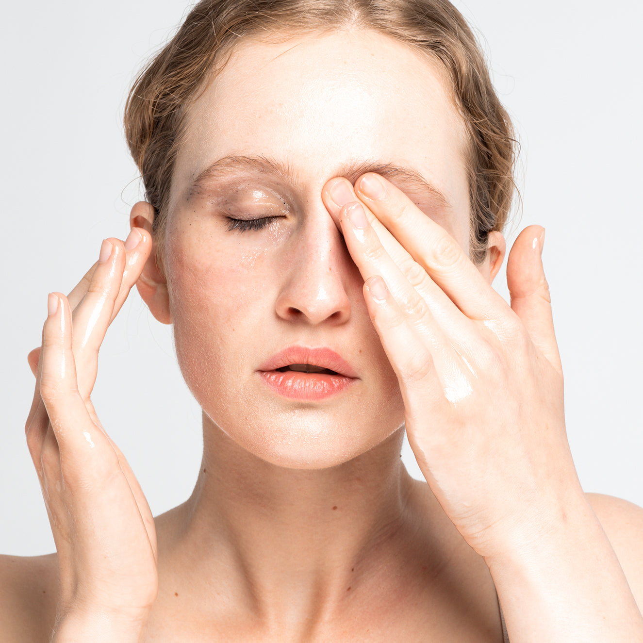 Frau reinigt ihr Gesicht mit geschlossenen Augen und feuchten Händen vor hellem Hintergrund.
