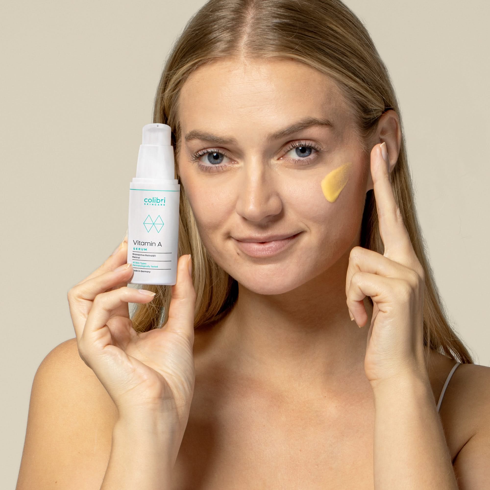 Frau hält ein Vitamin A Serum und zeigt auf ihr Gesicht, wo sie ein gelbes Hautpflegeprodukt aufgetragen hat.