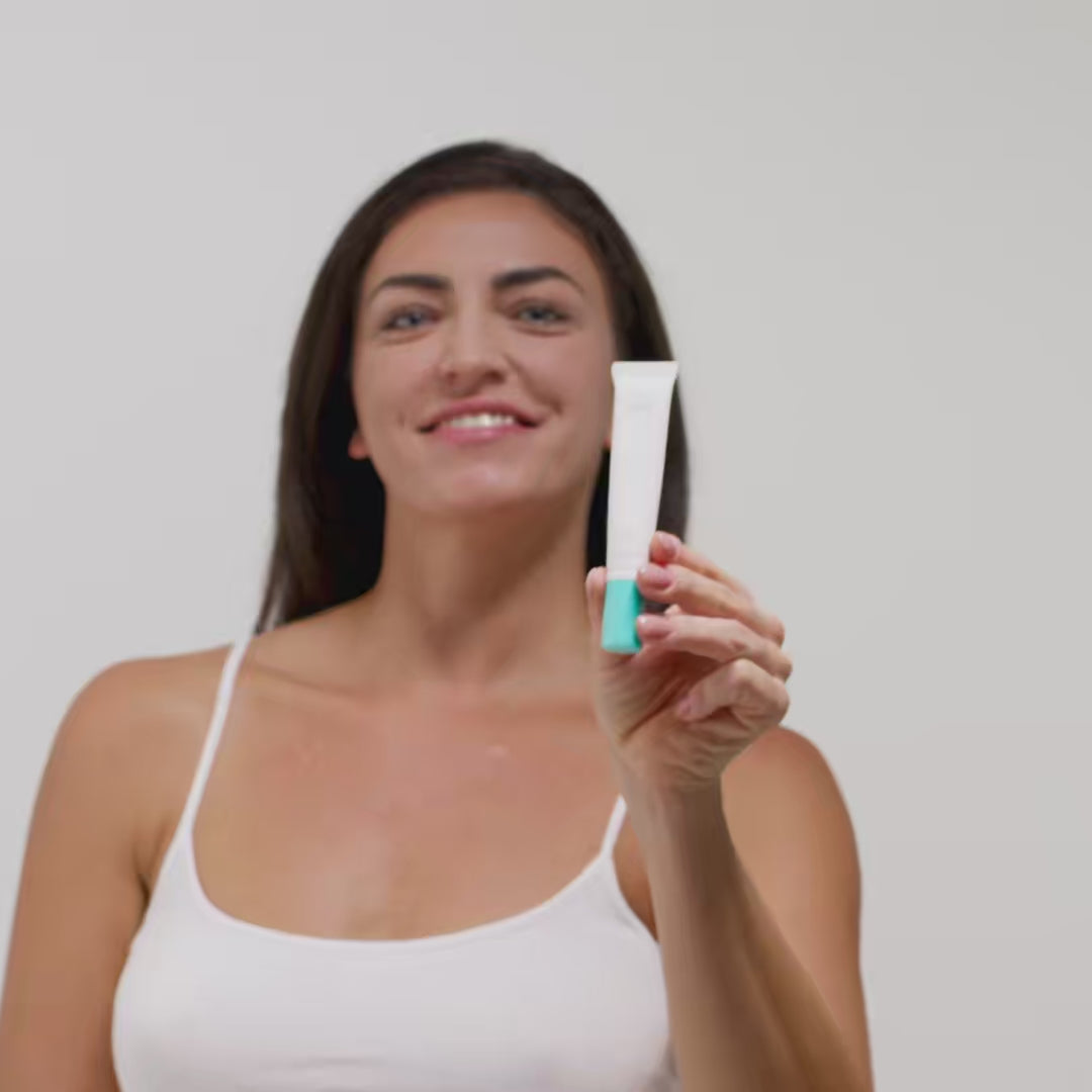 Eine Frau in einem weißen Top hält eine Tube mit einem türkisfarbenen Verschluss vor einem weißen Hintergrund.
