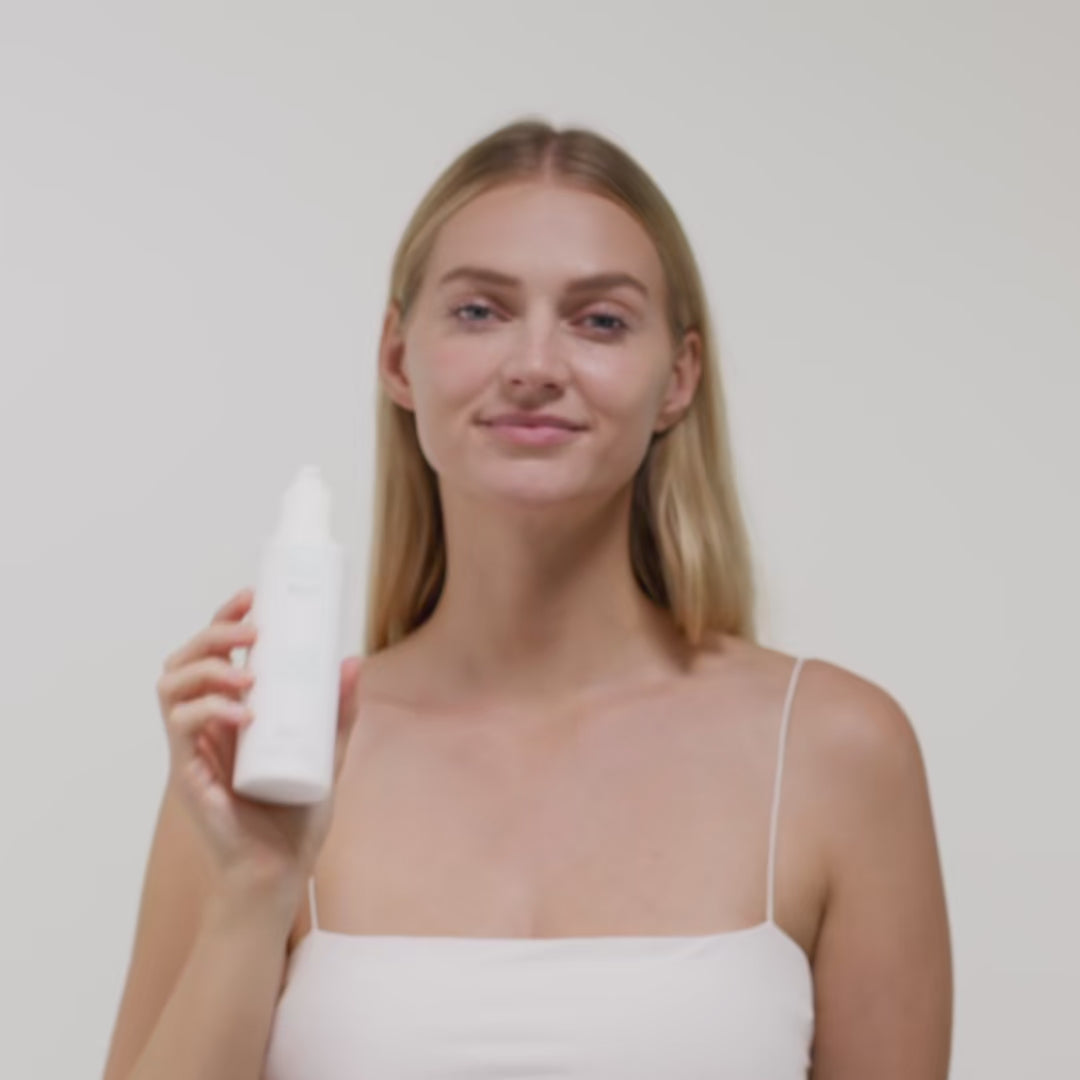 Frau mit langen Haaren hält eine weiße Flasche in der Hand, steht vor einem neutralen Hintergrund.