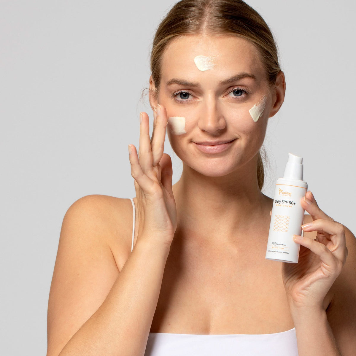 Eine Frau trägt Sonnencreme auf das Gesicht auf und hält eine Flasche mit der Aufschrift "Daily SPF 50+" in der Hand.